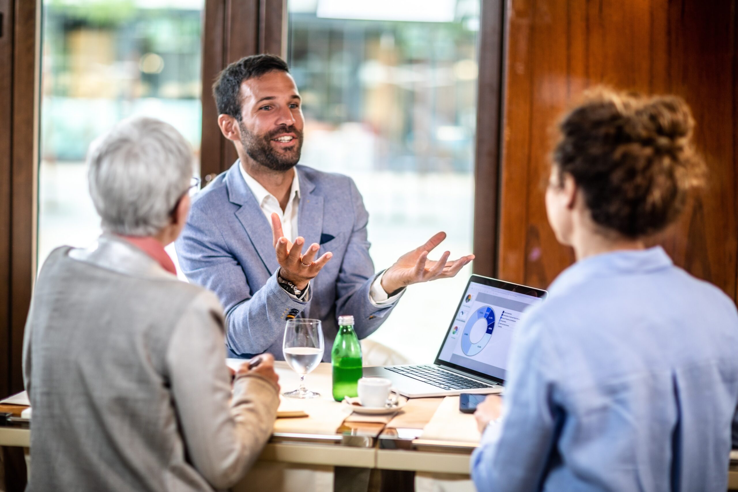 Accountant explaining financial report to two female co-founders in a restaurant. A laptop displaying a pie chart, a coffee cup, a water bottle and a glass are on the table.